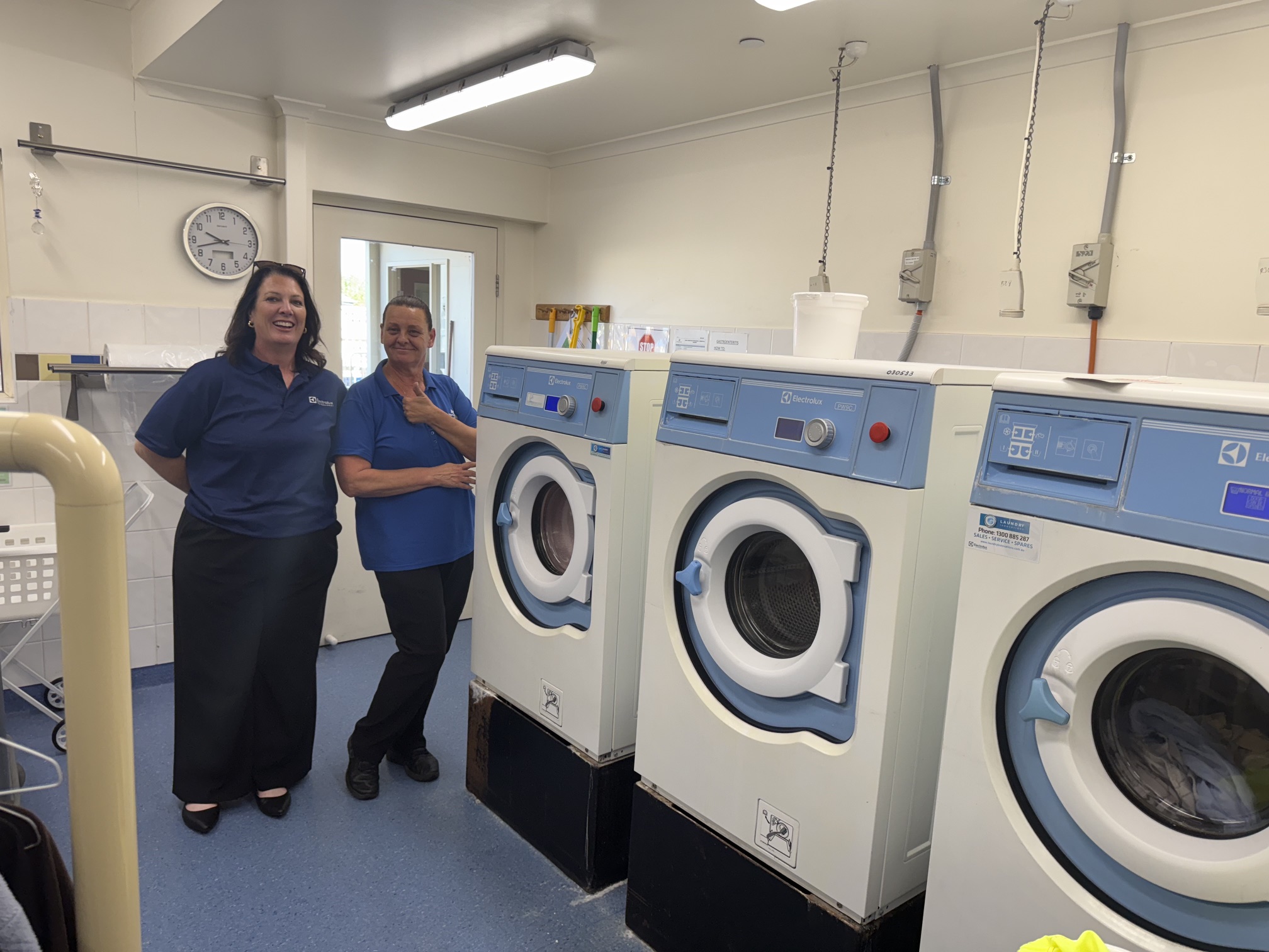 Two people standing beside installed washer extractors in an aged care laundry