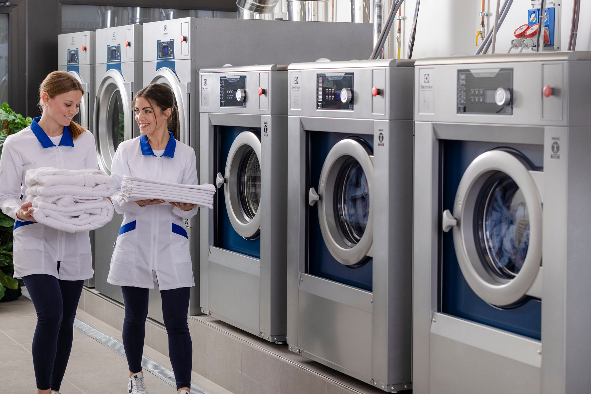 Two laundry staff holding folded white linens beside a row of Electrolux Professional commercial washer extractors